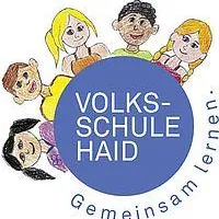 Das Bild zeigt eine Gruppe von Kindern um einen blauen Kreis herum, mit dem Text 'VOLKS-SCHULE HAID'. Die Kinder lächeln und scheinen Teil einer Schulgemeinschaft zu sein. Der Kreis hat einen blauen Hintergrund mit weißem Text.