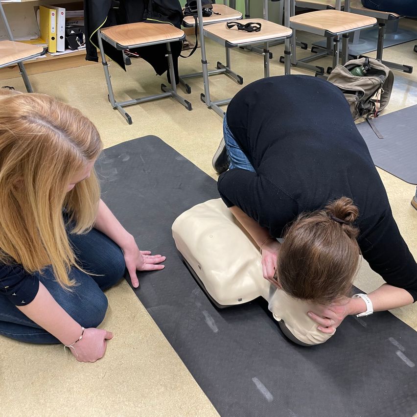 Zwei Frauen in einem Klassenzimmer, eine praktiziert CPR auf einem Dummies, während die andere genau zusieht. Hinter ihnen sind Tische gestapelt und ein Rucksack liegt auf dem Boden.