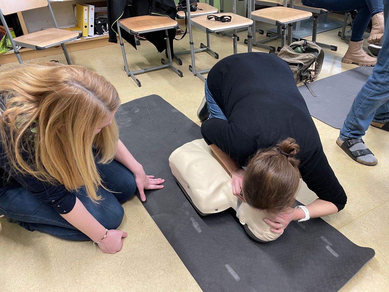 Zwei Frauen in einem Klassenzimmer, eine praktiziert CPR auf einem Dummies, während die andere genau zusieht. Hinter ihnen sind Tische gestapelt und ein Rucksack liegt auf dem Boden.