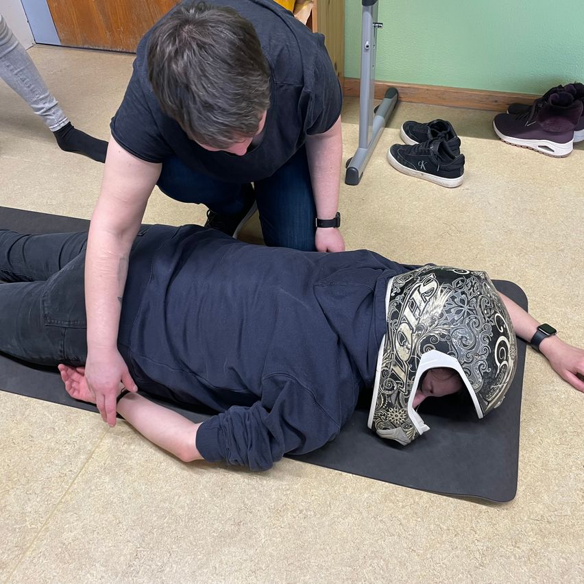 Eine Person, die auf einer Matte liegt, wird von einer knienden Person untersucht. Die liegende Person trägt einen Helm und eine Uhr. In der Nähe sind Schuhe und ein stehender Schreibtisch zu sehen.