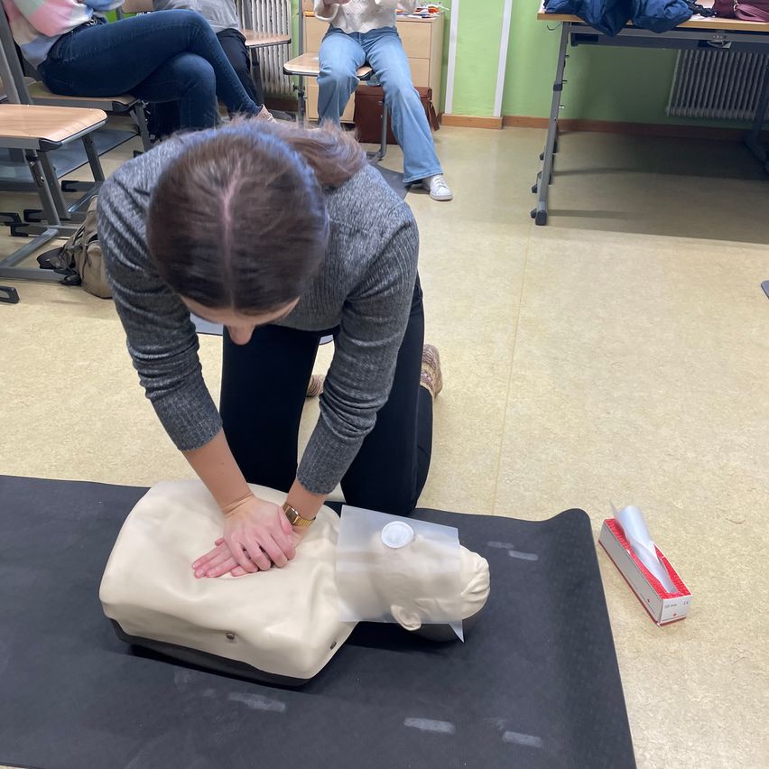 Eine Frau übt CPR an einem Dummies in einem Klassenzimmer. Eine Schachtel mit Latexhandschuhen ist in der Nähe.