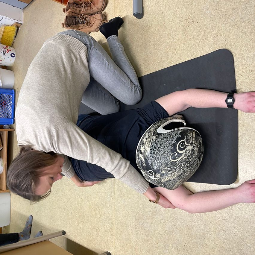 Eine Person liegt auf einer Yogamatte und führt eine Dehnübung durch, wobei eine andere Person von hinten hilft. Beide tragen Freizeitkleidung und die Person auf der Matte hält einen dekorativen Helm.