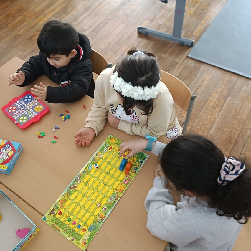 Drei Kinder sitzen um einen Tisch herum und spielen ein Brettspiel. Der Junge schaut auf ein kleines elektronisches Spiel. Das Mädchen in der Mitte hält einen Würfel. Das Mädchen auf der rechten Seite hat ein Band im Haar.