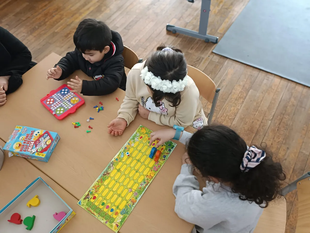 Drei Kinder sitzen um einen Tisch herum und spielen ein Brettspiel. Der Junge schaut auf ein kleines elektronisches Spiel. Das Mädchen in der Mitte hält einen Würfel. Das Mädchen auf der rechten Seite hat ein Band im Haar.