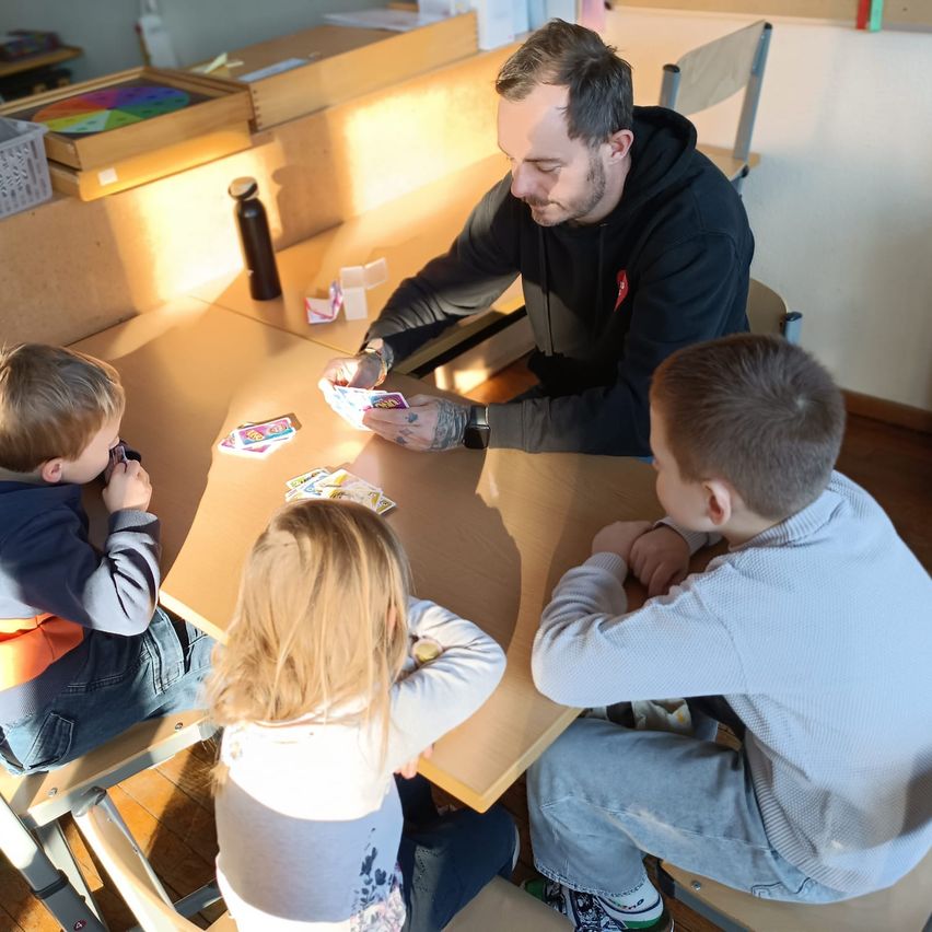 Ein Mann und drei Kinder sitzen um einen Tisch herum und spielen ein Kartenspiel. Der Mann hält eine Karte in der Hand. Die Kinder beobachten aufmerksam das Spiel.