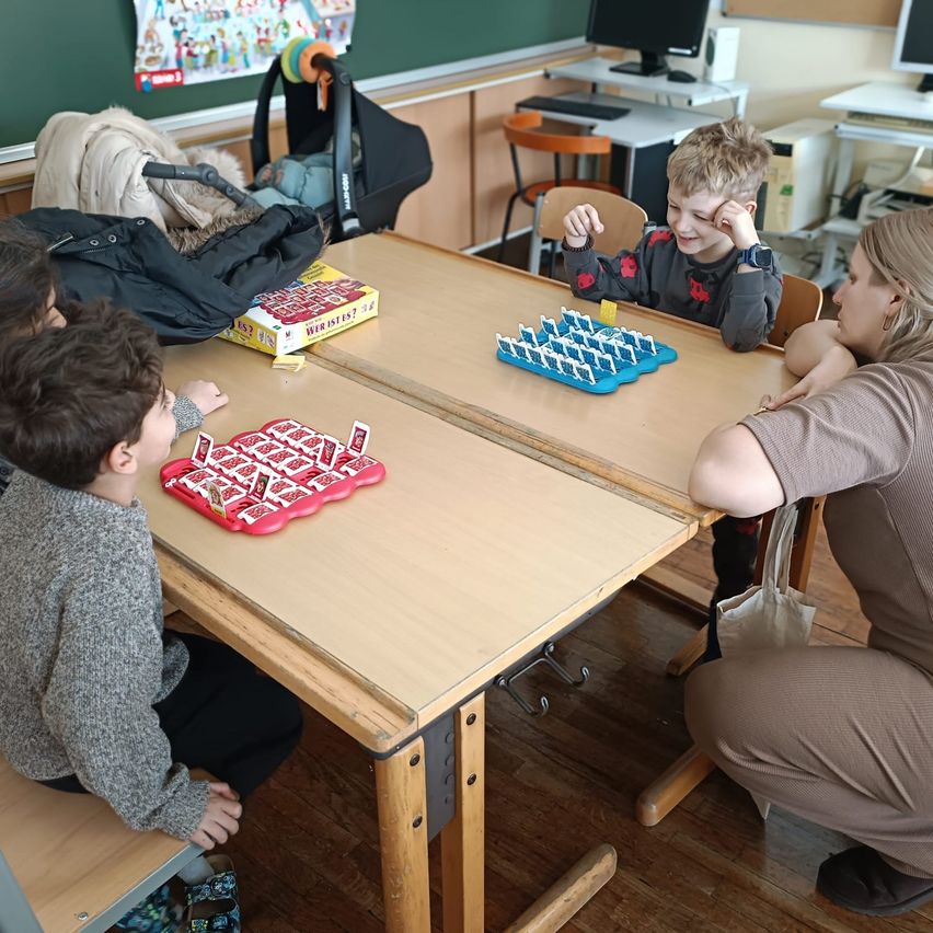 Eine Gruppe von Kindern spielt ein Brettspiel an einem Schreibtisch, eine Frau beobachtet. Ein Kinderwagen und ein Monitor sind im Hintergrund.