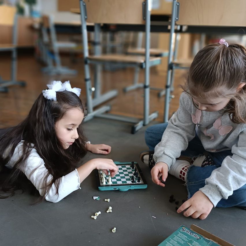 Zwei kleine Mädchen spielen Schach auf dem Boden in einem Klassenzimmer, mit verstreuten Schachfiguren um sie herum.
