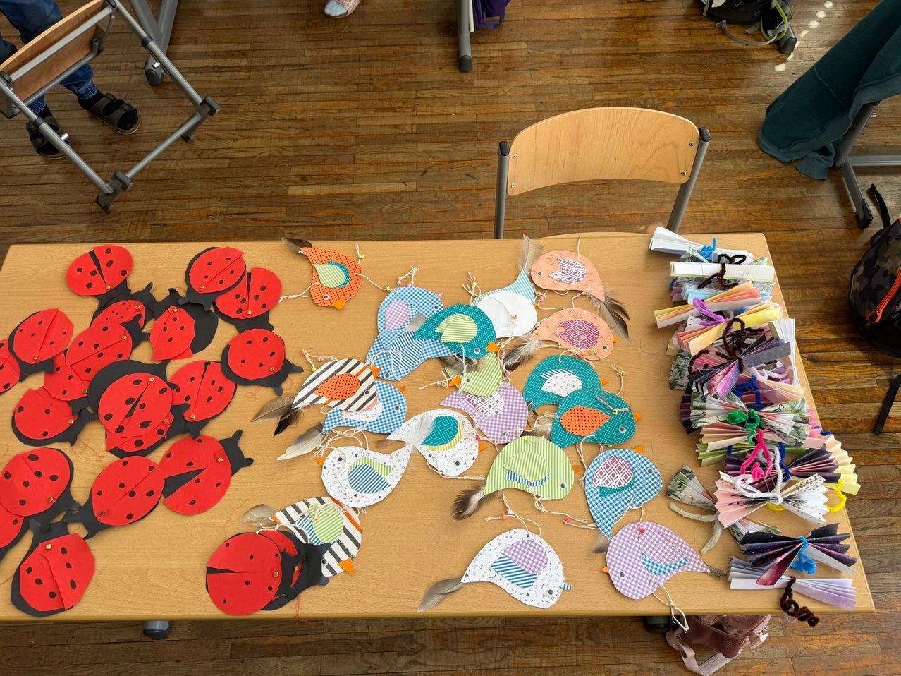 Ein Holztisch, der mit bunten Papierbasteleien, darunter Marienkäfer und Vögel, auf einem Holzboden bedeckt ist.