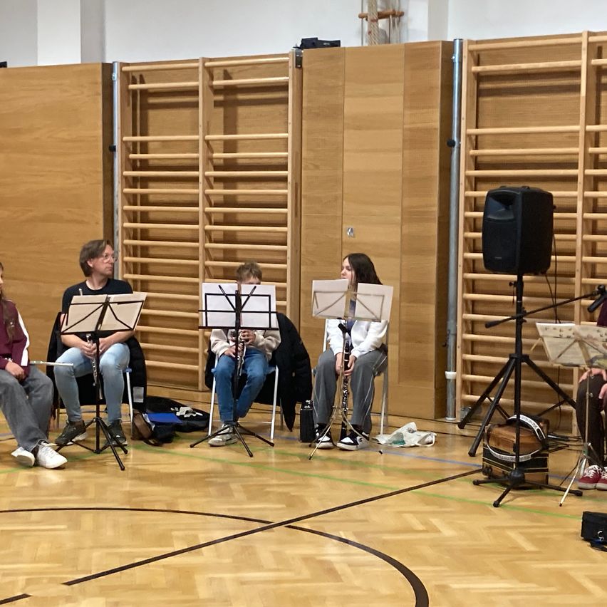 Eine Gruppe von Menschen in einem Saal übt Musik. Vier Personen sitzen auf Stühlen mit Notenständern vor ihnen. Sie spielen Flöten. Ein Lautsprecher steht auf einem Ständer rechts.