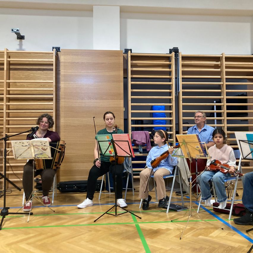 Eine Gruppe von Musikern, darunter eine erwachsene Frau und drei Kinder, üben Geige in einem Musikzimmer mit Holzpaneelen und Holzboden.