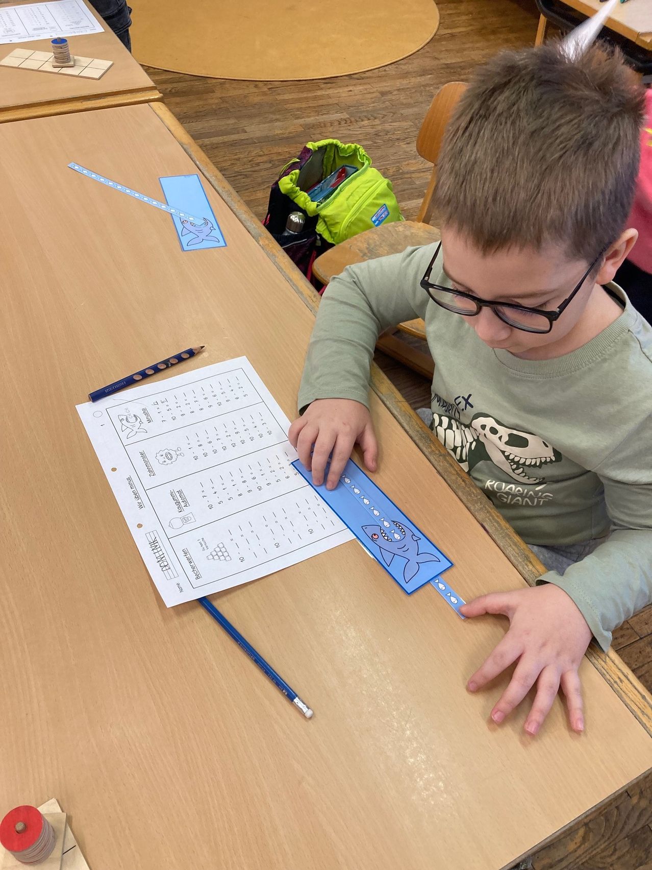 Ein Junge mit Brille sitzt an einem Schreibtisch und misst mit einem blauen Lineal ein Blatt mit mathematischen Aufgaben.