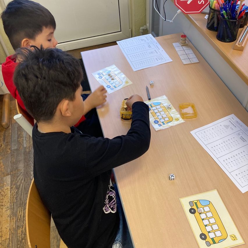 Zwei Jungen spielen ein Spiel auf einem Tisch mit Würfeln und einem Bus. Sie benutzen Stifte und Papiere.