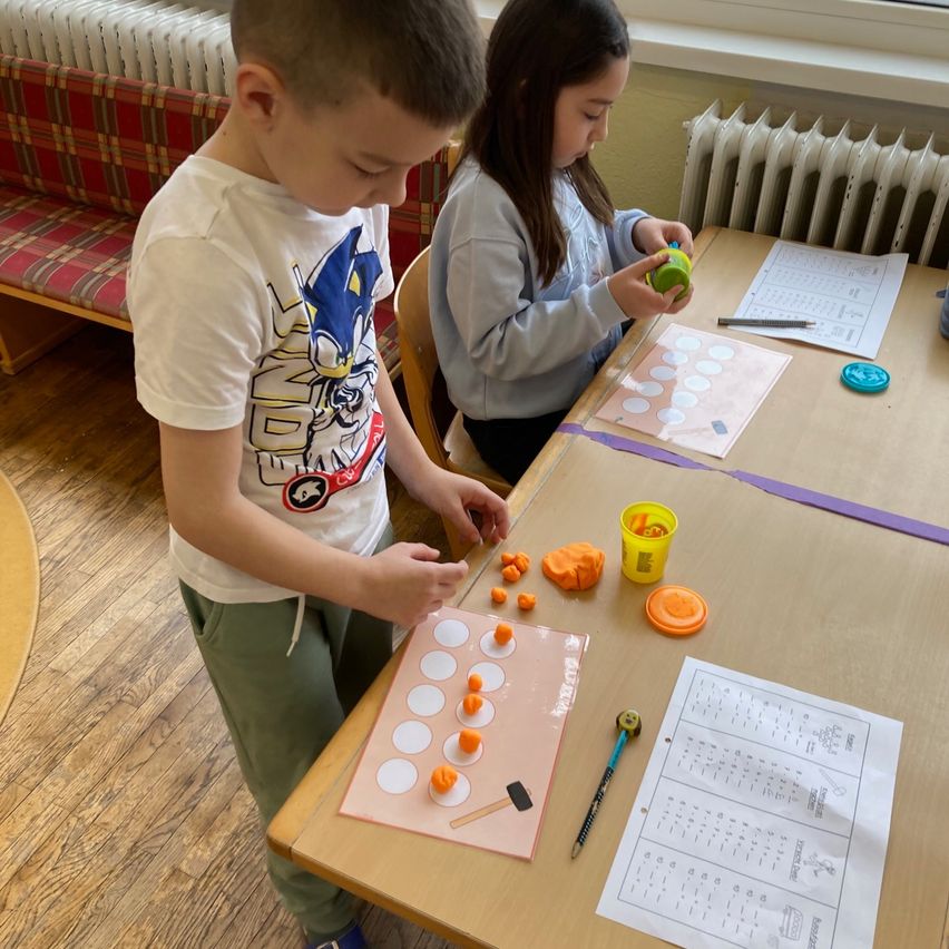 Ein Junge und ein Mädchen sitzen an einem Tisch und beschäftigen sich mit einem Bastelprojekt. Der Junge trägt ein weißes T-Shirt mit einem Comic-Charakter, und das Mädchen trägt ein hellblaues Langarmshirt. Verschiedene Bastelmaterialien liegen auf dem Tisch.