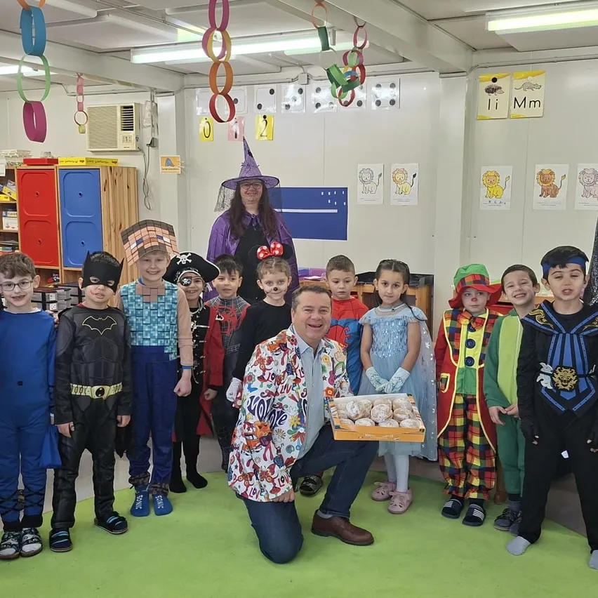 Eine Gruppe von Kindern in verschiedenen Kostümen steht in einem Klassenzimmer. Eine Frau in einer Hexenkostüm hält eine Tablett mit Donuts. Ein Mann hockt und hält das Tablett. Bunte Dekorationen hängen von der Decke.