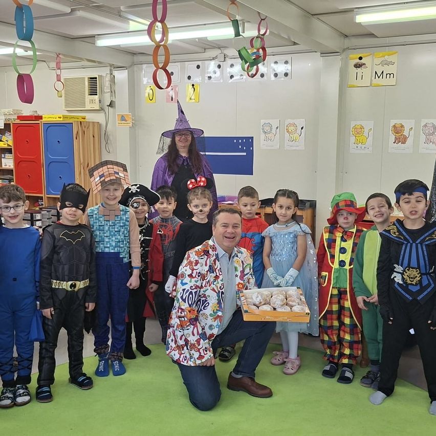 Eine Gruppe von Kindern in verschiedenen Kostümen steht in einem Klassenzimmer. Eine Frau in einer Hexenkostüm hält eine Tablett mit Donuts. Ein Mann hockt und hält das Tablett. Bunte Dekorationen hängen von der Decke.