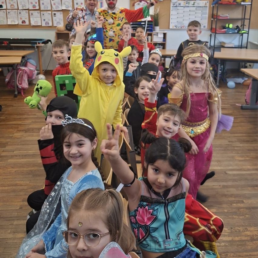 Gruppe von Kindern in Kostümen, die für ein Foto in einem Klassenzimmer posieren, mit Erwachsenen, Tischen, Regalen und einer Ankündigungstafel im Hintergrund.