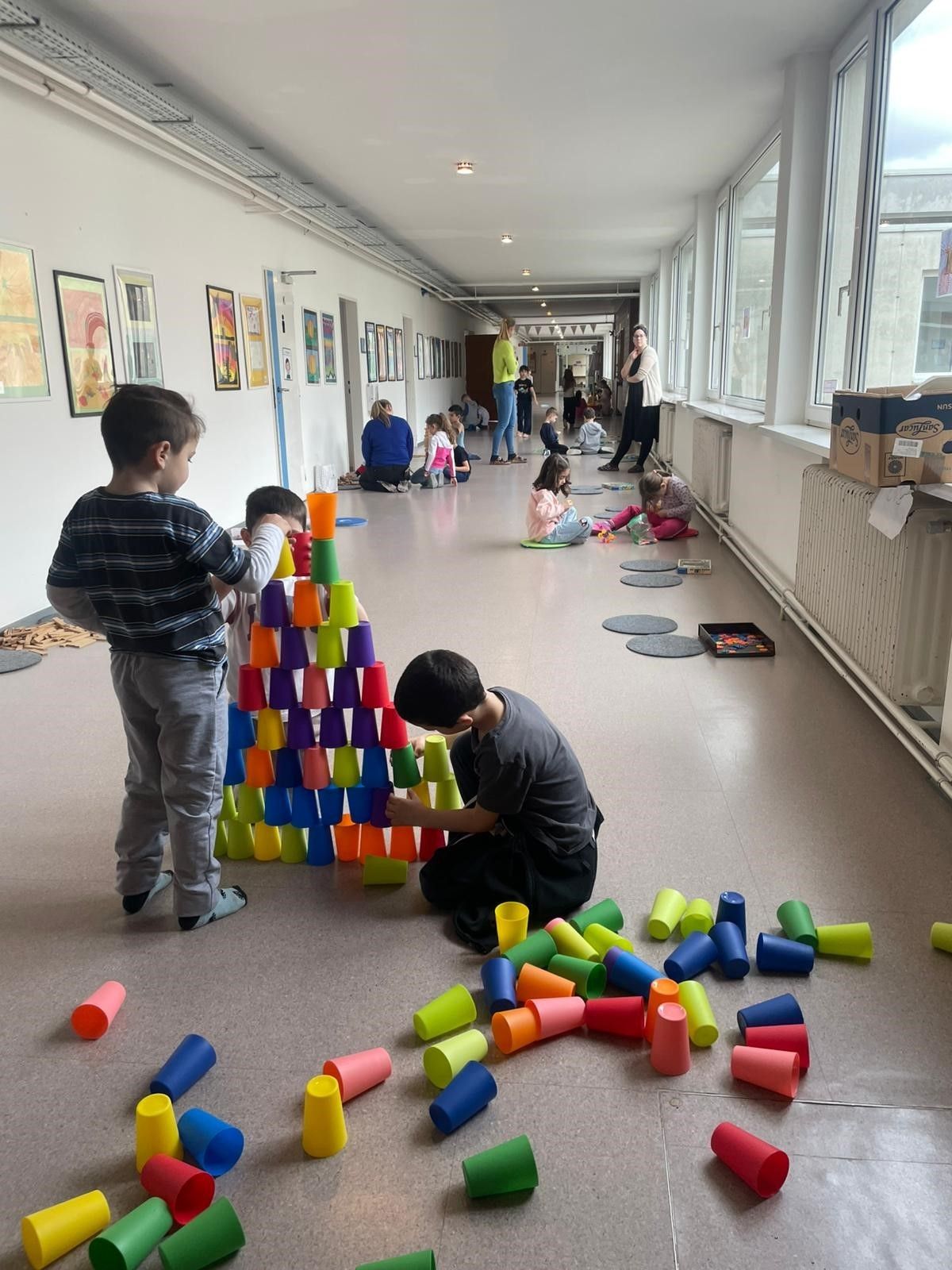 Kinder spielen mit Bechern in einem Flur. Ein Junge stapelt Becher, während ein anderer kniet und sie umwirft. Erwachsene beobachten von der Seite.