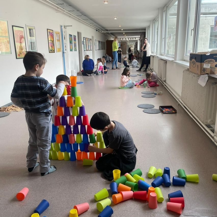 Kinder spielen mit Bechern in einem Flur. Ein Junge stapelt Becher, während ein anderer kniet und sie umwirft. Erwachsene beobachten von der Seite.