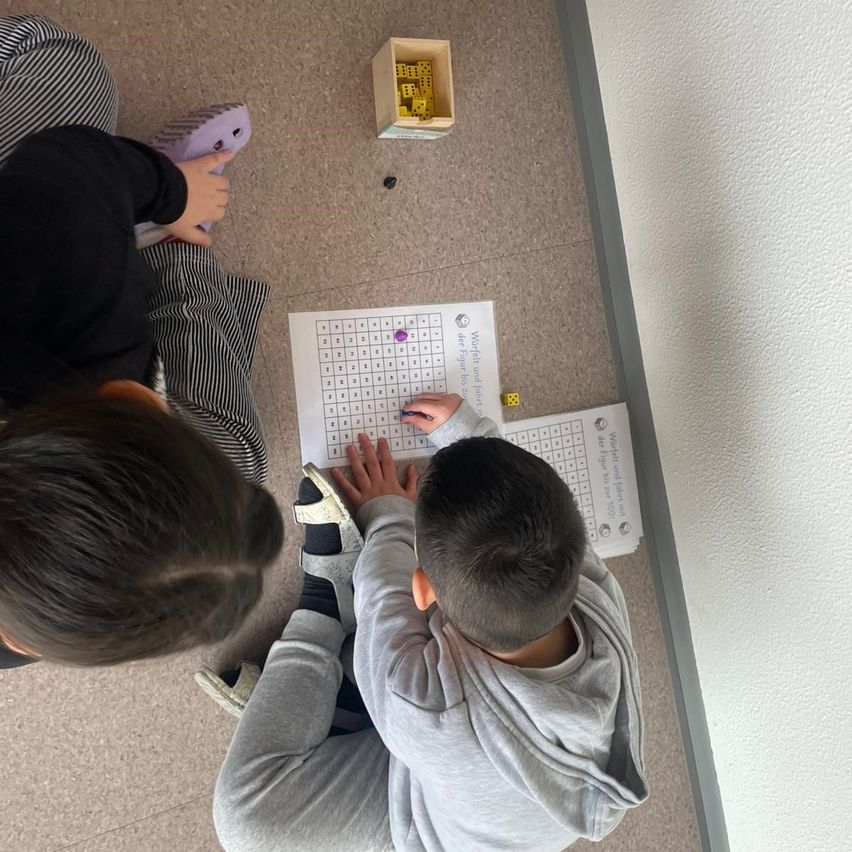 Zwei Kinder sitzen auf dem Boden, eines hält einen Würfel und das andere einen Stift. Sie spielen ein Spiel auf einem Blatt Papier.