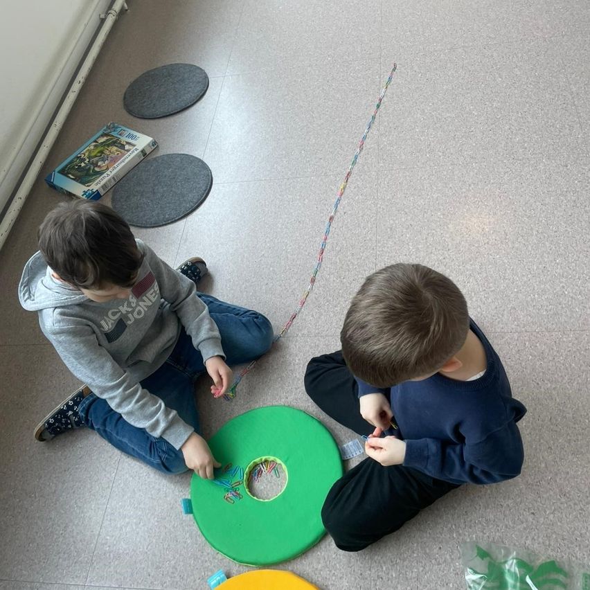 Zwei Jungen sitzen auf dem Boden und spielen mit einem grünen Objekt und einer bunten Schnur. Sie betrachten es aufmerksam. Auf dem Boden liegen drei graue Kreise und ein Puzzle.