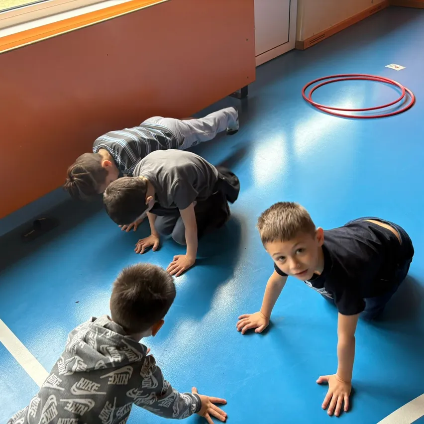 Vier junge Jungen machen Liegestütze auf einem blauen Boden mit einem roten Hula-Hoop im Hintergrund. Die Jungen tragen Freizeitkleidung, und einer hat ein Nike-Logo auf seiner Kapuzenjacke.
