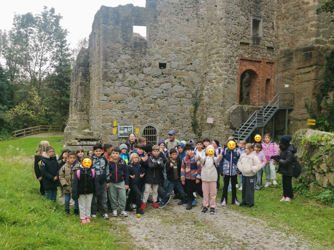 Eine Gruppe von Kindern steht vor einem alten Steingebäude. Sie tragen Winterkleidung und einige haben Emojis auf den Gesichtern. Im Hintergrund sind Bäume und eine Treppe sichtbar.