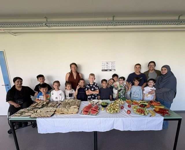 Eine Gruppe von Menschen, einschließlich Erwachsener und Kindern, steht hinter einem Tisch mit verschiedenen Essensartikeln. Sie scheinen für ein Foto zu posieren.