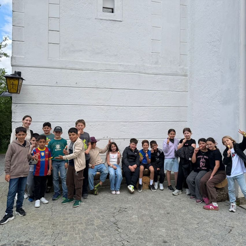 Eine Gruppe von Kindern und Jugendlichen posiert für ein Foto vor einem weißen Gebäude. Einige sitzen auf Baumstümpfen, während andere stehen, und alle haben Eis am Stiel.