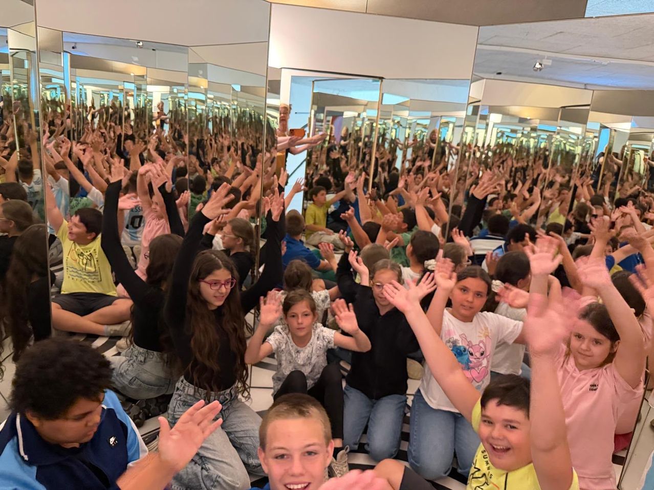 Ein Klassenzimmer voller Kinder, die ihre Hände in einem verspiegelten Raum heben, einige tragen Brillen, Turnschuhe und ein gelbes T-Shirt mit einem rosa Herzen-Design.