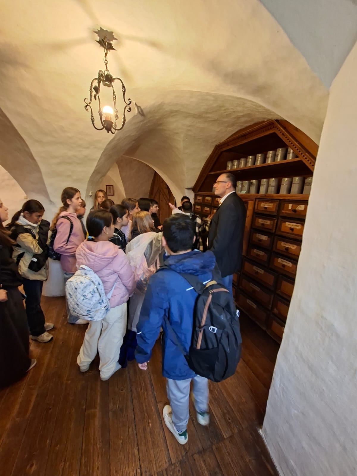 Eine Gruppe von Schülern schaut sich einen Holzschrank mit vielen Schubladen an, ein Mann spricht mit ihnen in einem Raum mit Holzböden und gewölbten Decken.