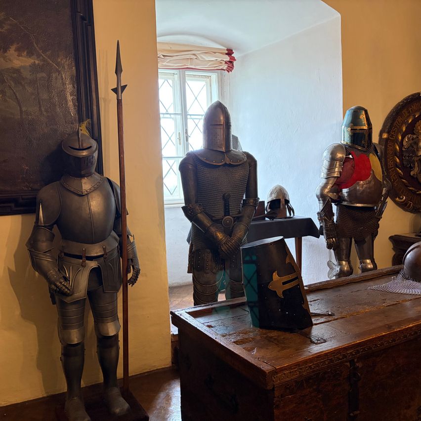 Gepanzerte Ritter stehen in einem Raum mit Holzmöbeln. Ein Helm und eine Lanze sind in der Nähe. Ein Fenster und ein Gemälde an der Wand sind sichtbar.