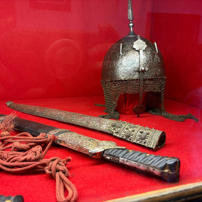 Ein alter Metallhelm und drei kunstvoll verzierte Schwerter sind auf einem roten Tuch in einer Glasvitrine ausgestellt. Die Schwerter haben Quasten und dekorative Gravuren.