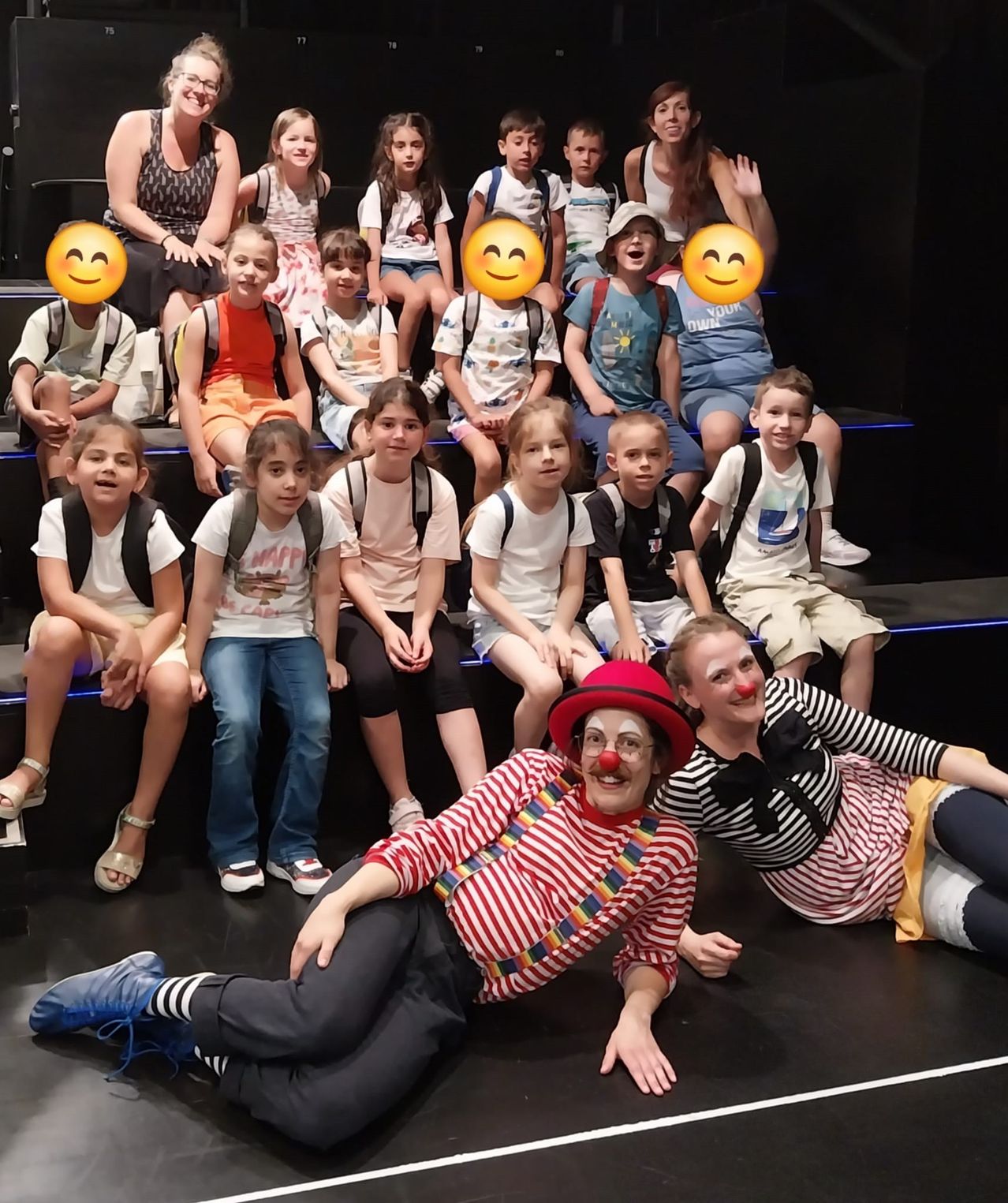 Eine Gruppe von Kindern und Erwachsenen steht auf einer Bühne. Die Kinder tragen Rucksäcke und lächeln. Zwei Clowns in farbenfrohen Kostümen liegen auf dem Boden.