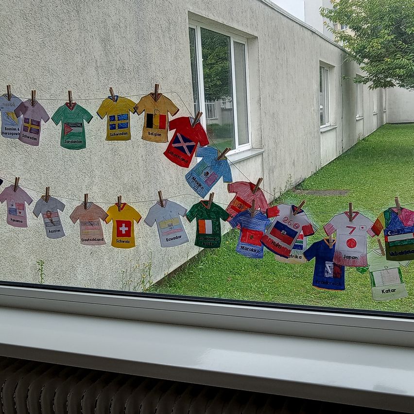 Eine Reihe bunter bemalter Fußballtrikots, die an einer Wäscheleine vor einem Gebäude mit Fenster und einem Baum im Hintergrund hängen.