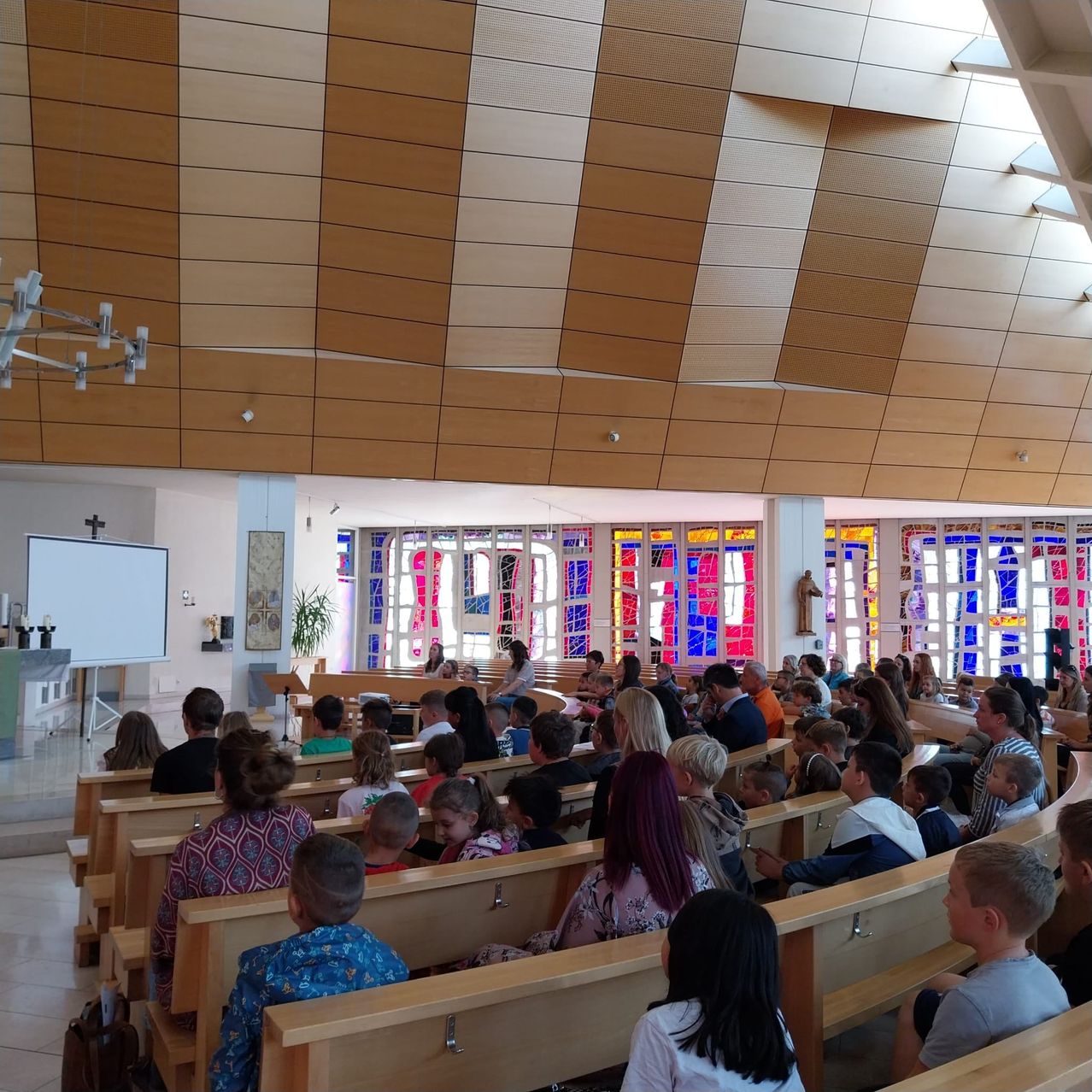 Eine Gemeinde füllt Holzbänke in einer Kirche, die auf eine Bühne mit einer Tafel blicken. Die Decke hat ein gestreiftes Design. Bunte Buntglasfenster werfen leuchtendes Licht.