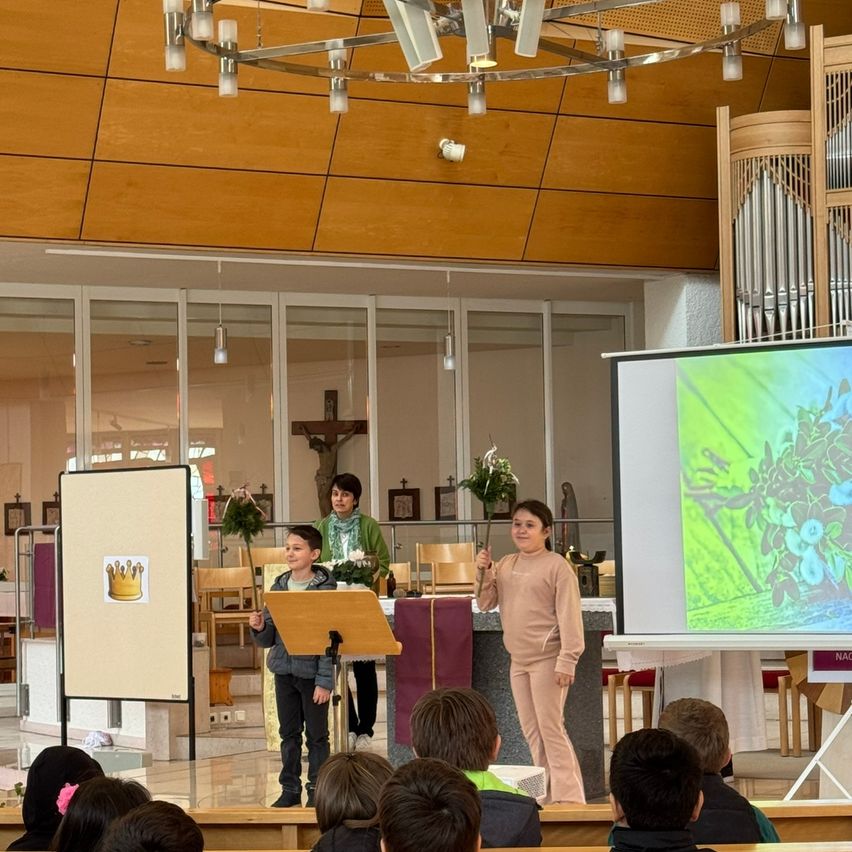 In einer Kirche findet eine Präsentation statt, bei der zwei Kinder vor einer Leinwand stehen. Eines hält ein Mikrofon, während das andere eine Krone auf einem Schild hat. Dahinter hält eine Frau ein Mikrofon und Blumen.