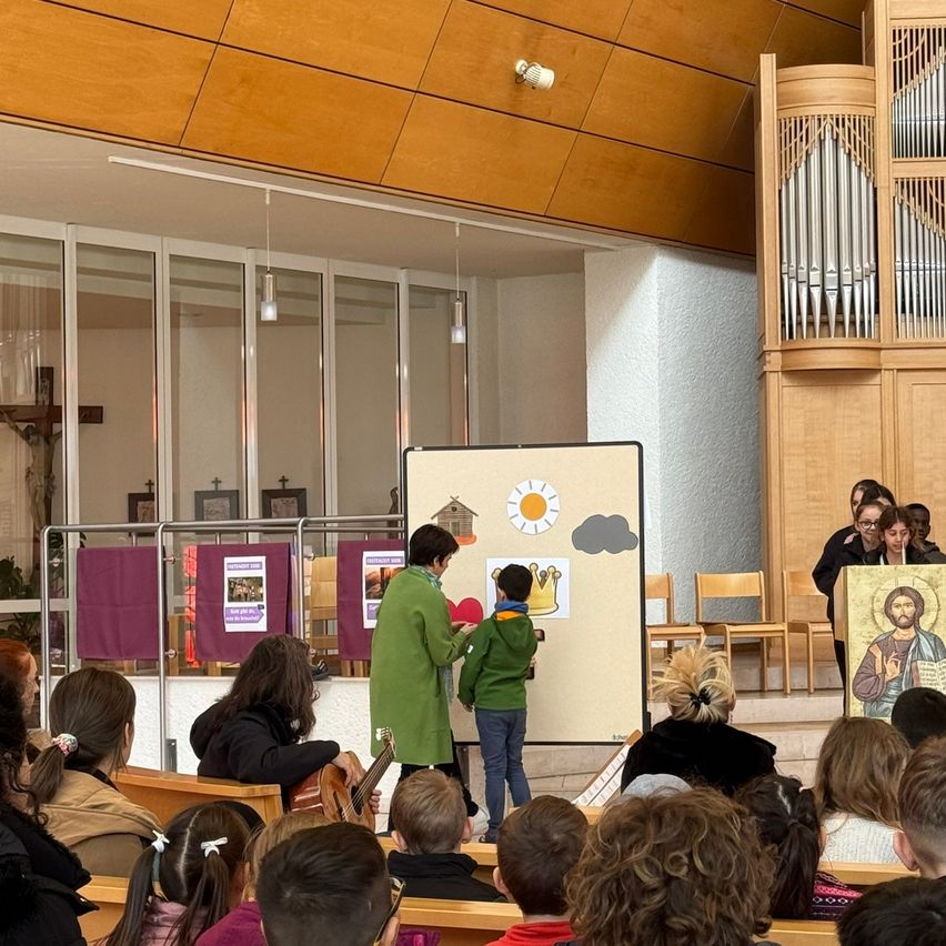 Eine Kirchenveranstaltung mit einem Publikum, das auf Bänken sitzt. Eine Person in Grün hilft einem Kind vor einer Tafel mit Zeichnungen. Eine Orgel befindet sich auf der rechten Seite und ein hölzerne Kreuz auf der linken.
