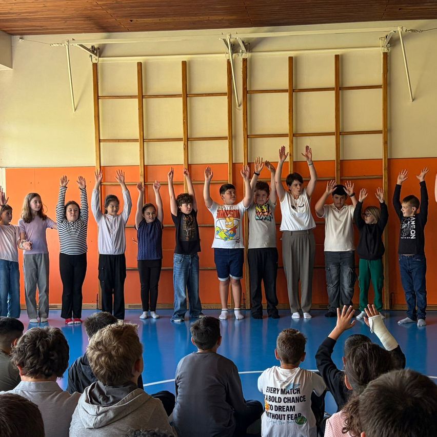 Eine Gruppe von Kindern und Jugendlichen steht mit erhobenen Händen in einer Reihe in einer Turnhalle. Einige von ihnen tragen Brillen, Sneaker und Armbänder. Sie stehen vor Holzleitern und einer orangefarbenen Wand. Einige Kinder sitzen vor ihnen auf dem Boden.