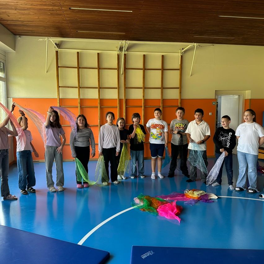 Eine Gruppe von Kindern steht in einer Turnhalle mit blauem Boden. Einige halten bunte Bänder. Rechts ist eine weiße Tür, und an der Wand sind Holzleitern befestigt.