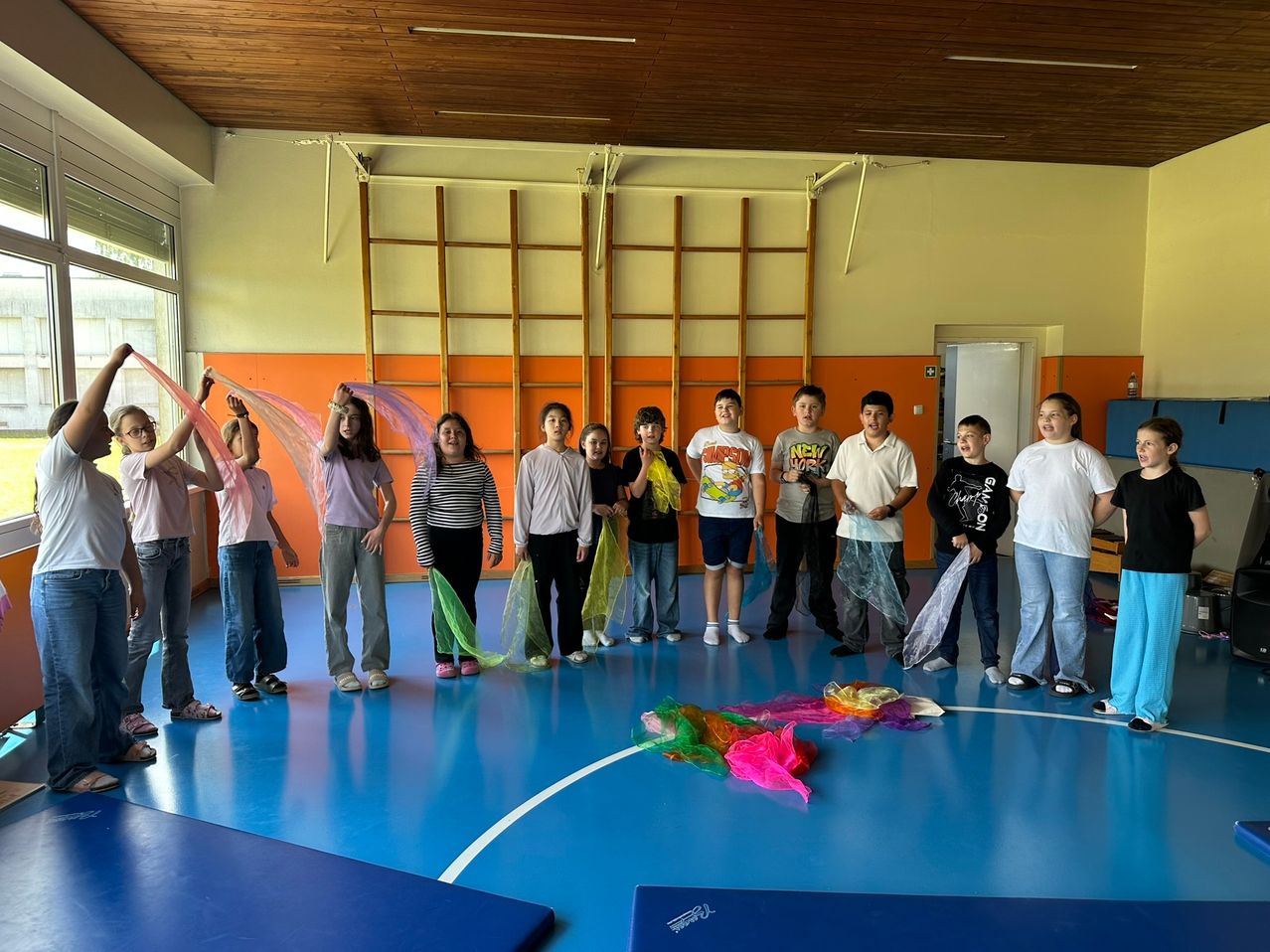Eine Gruppe von Kindern steht in einer Turnhalle mit blauem Boden. Einige halten bunte Bänder. Rechts ist eine weiße Tür, und an der Wand sind Holzleitern befestigt.