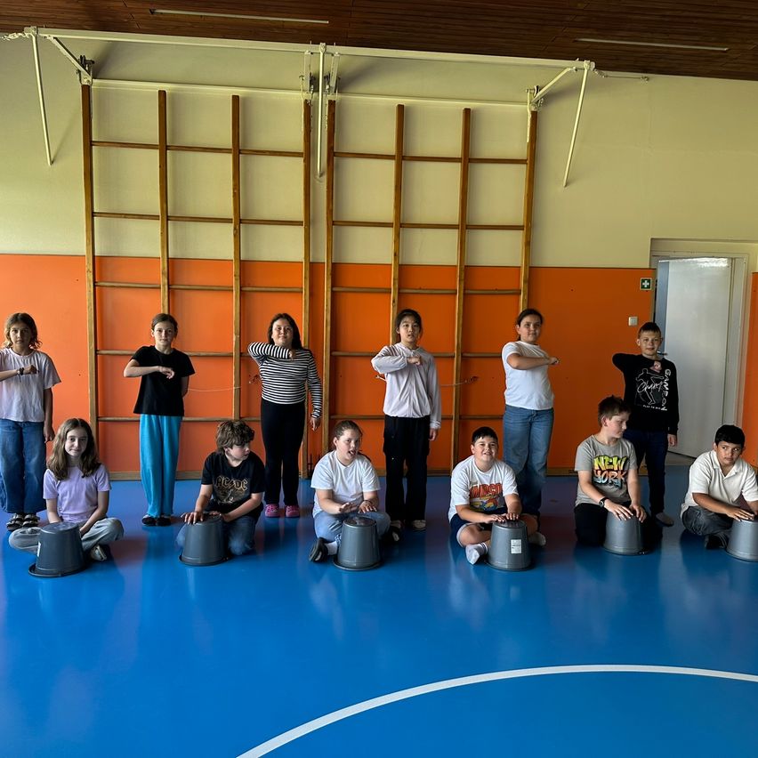 Mehrere Kinder sitzen mit Trommeln auf dem Boden, mit drei Holzleitern an der Wand. Sie scheinen für eine Aufführung zu üben.