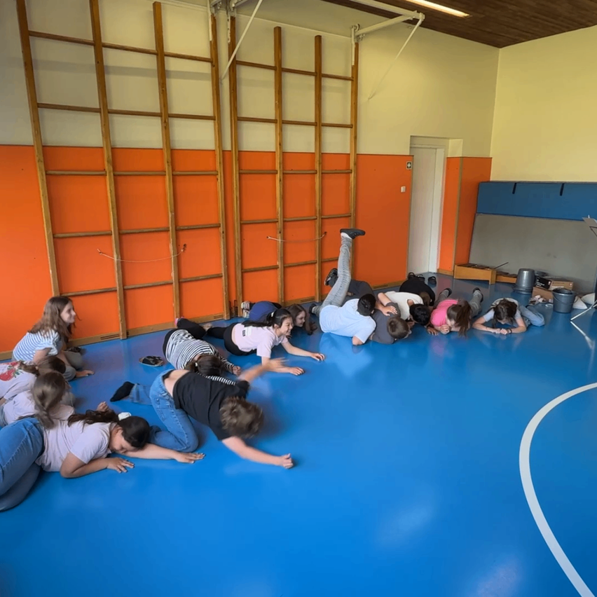 Eine Gruppe von Kindern liegt auf dem blauen Boden einer Turnhalle. Einige tragen Turnschuhe und einer hat einen Schuh an. Drei hölzerne Leitern sind an der orangefarbenen Wand befestigt.