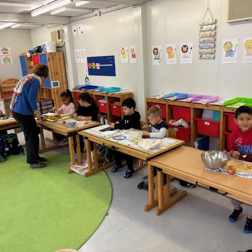 Eine Gruppe von Kindern sitzt an Tischen in einem Klassenzimmer, eine Lehrerin steht in der Nähe. Sie sind mit einer Aktivität beschäftigt, wahrscheinlich bereiten sie Essen vor.