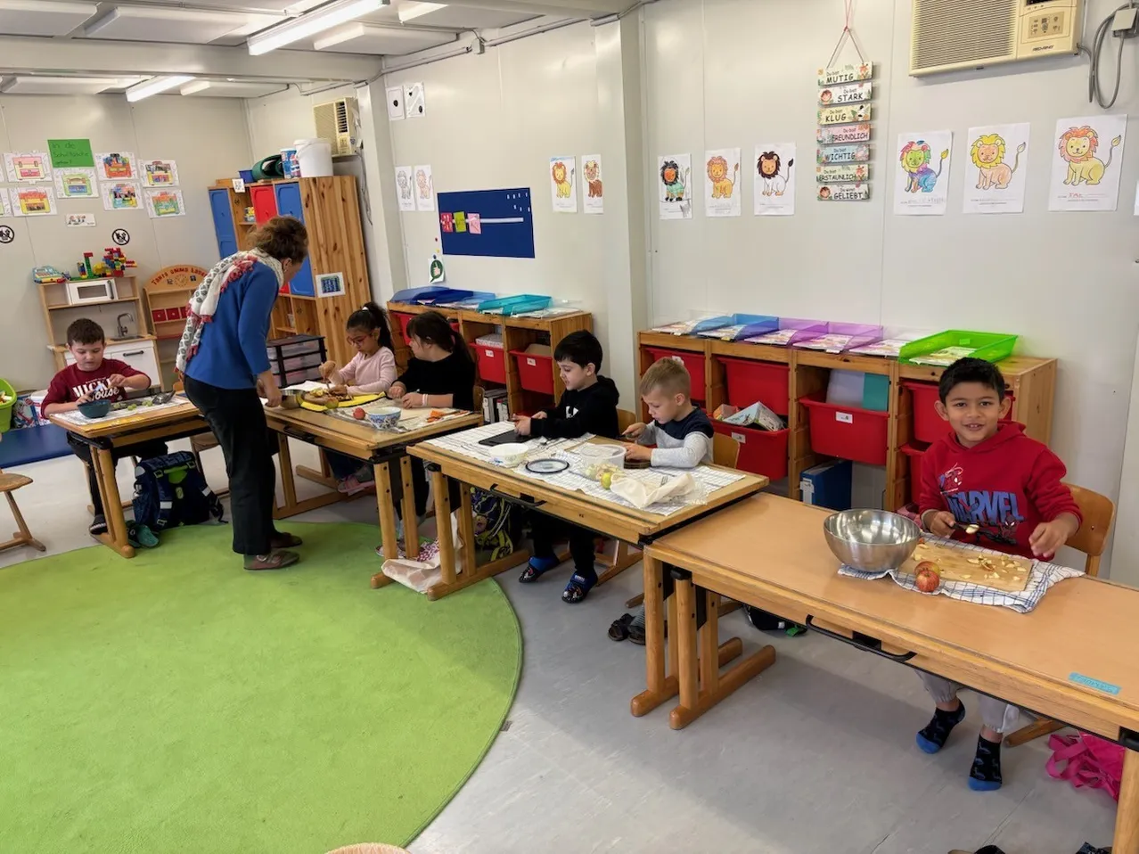 Eine Gruppe von Kindern sitzt an Tischen in einem Klassenzimmer, eine Lehrerin steht in der Nähe. Sie sind mit einer Aktivität beschäftigt, wahrscheinlich bereiten sie Essen vor.