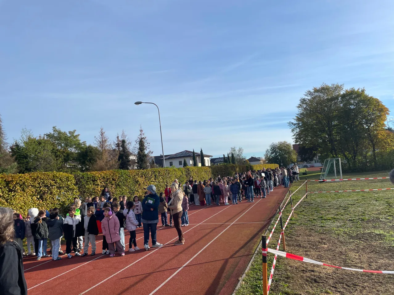 Eine Gruppe von Menschen, wahrscheinlich Schüler, stehen in einer Reihe auf einer roten Laufbahn, möglicherweise für ein Rennen. Die Laufbahn ist von einer Hecke umgeben, und es gibt eine Straßenlaterne an der Seite. In der Ferne befindet sich ein Fußballtor und Bäume.
