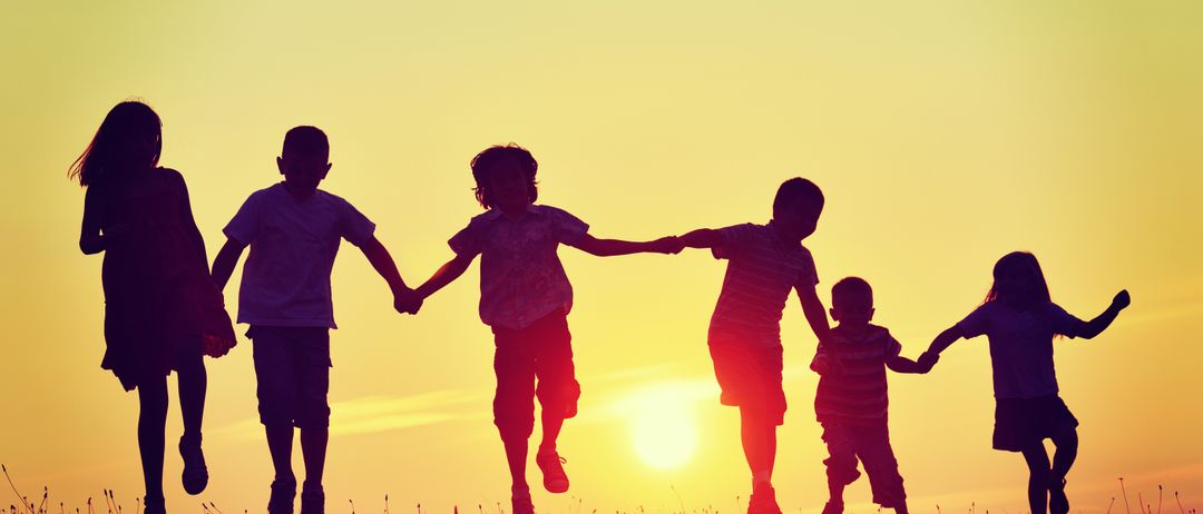 Fünf Kinder laufen Hand in Hand auf einem Feld mit dem untergehenden Sonnen im Hintergrund.