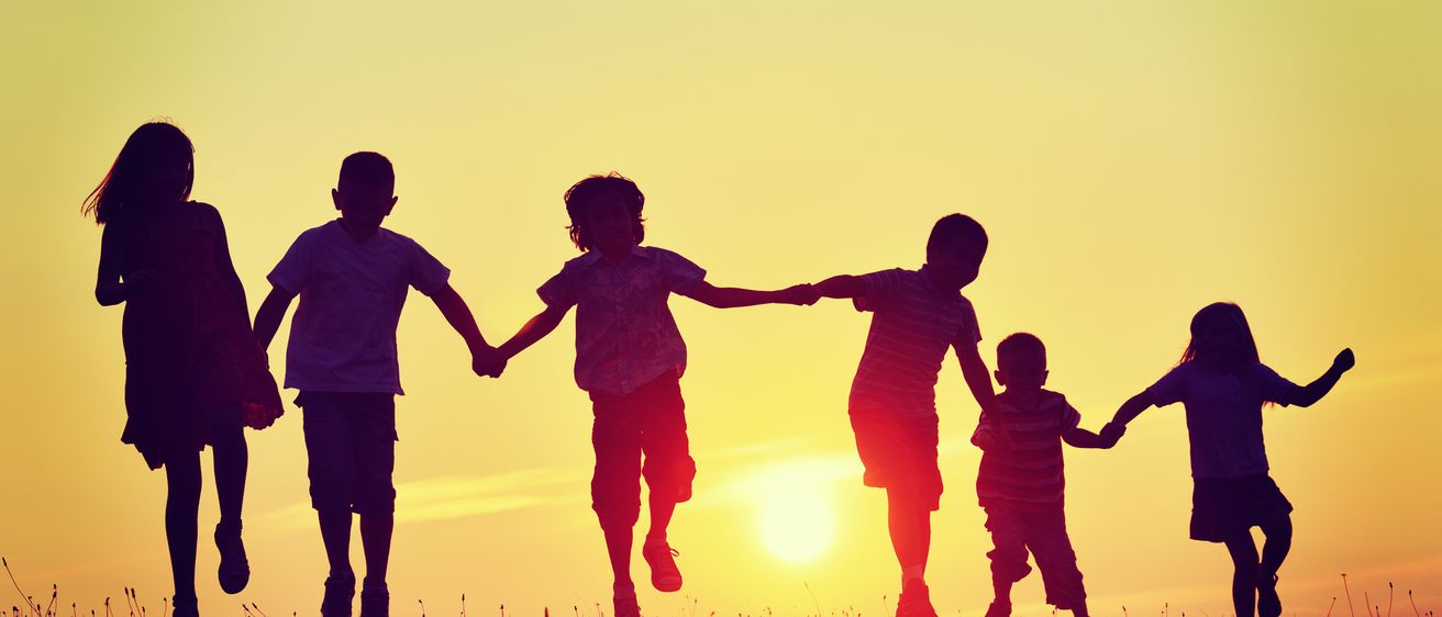 Fünf Kinder laufen Hand in Hand auf einem Feld mit dem untergehenden Sonnen im Hintergrund.