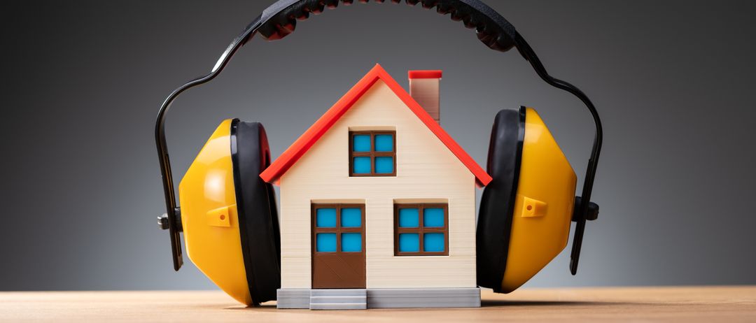 Ein kleines Hausmodell mit rotem Dach und blauen Fenstern steht auf einem Tisch. Es ist von gelben Ohrschützern umgeben.