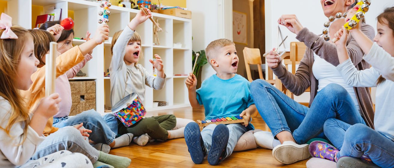 Mehrere Kinder sitzen in einem Raum auf dem Boden. Eine Frau spielt mit ihnen ein Musikinstrument. Die Kinder halten Instrumente und spielen mit.