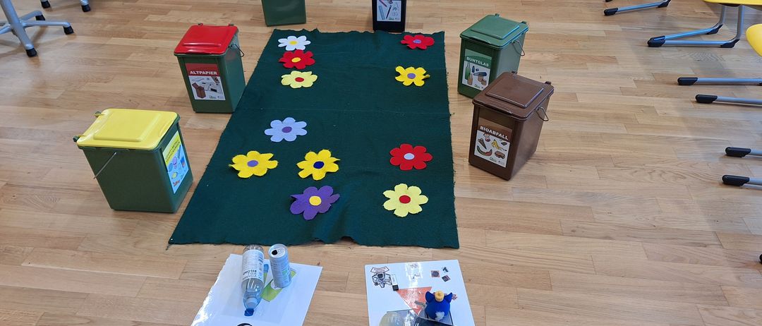 Ein grüner Teppich mit Blumen darauf liegt auf einem Holzboden, mit mehreren Recycling-Behältern darum. Links sind zwei Papiere mit Recycling-Symbolen. Rechts steht ein brauner Kasten mit der Aufschrift Bioabfall.
